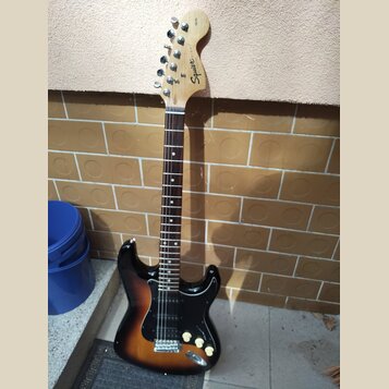 Elektrická kytara s trupem v barevnosti sunburst a černým pickguardem. Na hlavě hmatníku je vidět nápis Squier by Fender. Nástroj má tři snímače typu…