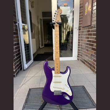 Fialová elektrická kytara s bílým pickguardem a javorovým hmatníkem. Na hlavě je viditelné logo Fender. Nástroj je umístěn na černém stojanu.