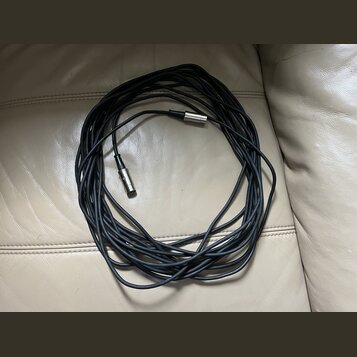 Černý kabel s kovovými konektory na obou koncích. Kabel je svinutý do kruhu a položený na světlém koženém povrchu. Na konektorech nejsou viditelné žá…