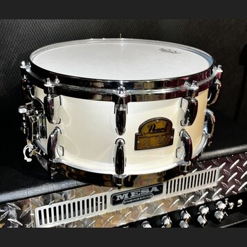 Bílá buben (snare) s chromovanými obručmi a ladicími šrouby. Na korpusu je zlatý oválný štít s nápisem Pearl a pod ním text SIGNATURE SERIES. Buben s…