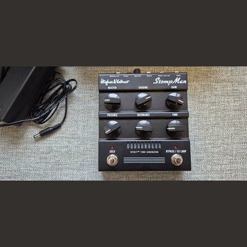 Kytarový efekt StompMan značky Hughes & Kettner v černé barvě. Na horní straně jsou nápisy Speaker out, Power, Solo Vol., Return, Send / Line out a I…