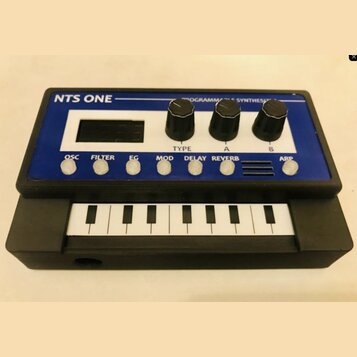 Černý kompaktní syntetizátor s modrým horním panelem a malou klaviaturou. Na horní části je bílý nápis NTS ONE a text PROGRAMMABLE SYNTHESIZER. Panel…
