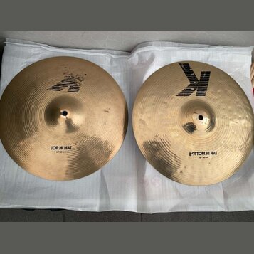 Dva mosazné činely typu hi-hat položené na bílé podložce. Horní činel má nápis TOP HI HAT a logo Zildjian. Spodní činel má nápis BOTTOM HI HAT a výra…