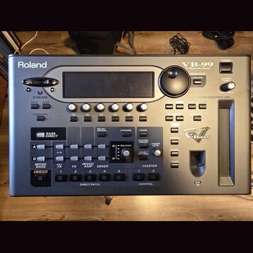Šedý multifunkční procesor pro basovou kytaru značky Roland, model VB-99. Zařízení obsahuje displej, množství tlačítek, posuvníky a ovládací knoflíky…
