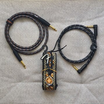 Na fotografii jsou dva instrumentální kabely a jeden kytarový popruh. Levý kabel je šedý s oranžovými proužky a nápisy Roland a RIO-05. Pravý kabel j…
