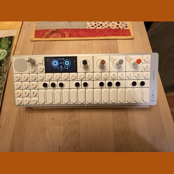 Bílý kompaktní synthesizer s klávesou a ovládacími prvky. Na pravém boku je svisle napsáno OP-1 field. Zařízení obsahuje displej, čtyři barevné otočn…
