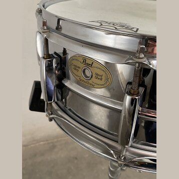 Detailní pohled na kovový snare drum stříbrné barvy s chromovaným obručováním. Na korpusu je oválný zlatý štítek s nápisy Pearl, Custom Alloy, Steel…