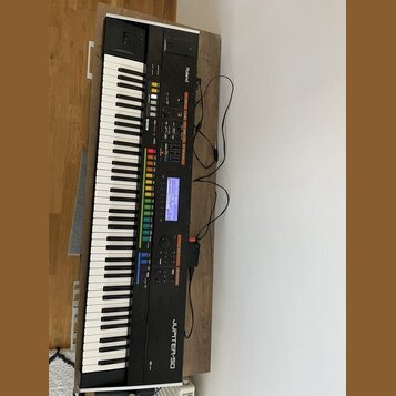 Černý digitální syntezátor s klaviaturou, umístěný na dřevěném stole. Na přístroji jsou viditelná loga a nápisy Roland a JUPITER-80. Ovládací panel o…