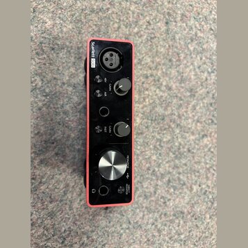 Červené audio rozhraní s nápisem Scarlett Solo. Na přední straně jsou dva otočné knoflíky GAIN, jeden velký knoflík MONITOR a tlačítka 48V, AIR, INST…