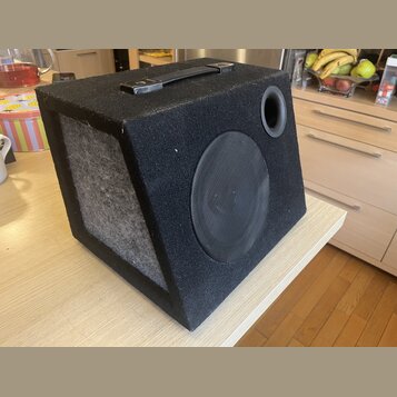 Černý kostkový subwoofer potažený texturovaným materiálem s madlem na horní straně. Přední strana obsahuje kruhový reproduktor krytý černou síťkou a…