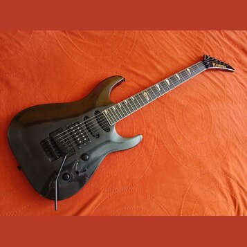Černá elektrická kytara s tmavým hnedým hmatníkem a špičatou hlavou. Na hlavě je žlutý nápis KRAGLER. Nástroj má dva humbuckery, jeden jednosmykový s…