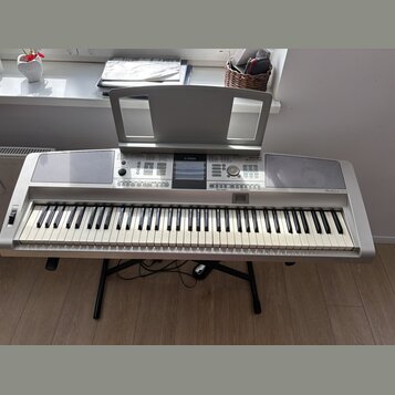 Stříbrný digitální klavír s černými a bílými klávesami, umístěný na černém X-stojanu. Na horním panelu je uprostřed logo YAMAHA a vpravo nápis Clavin…