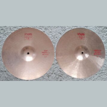 Dva mosazné činely typu hi-hat položené vedle sebe na bílém koberci. Oba mají červené nápisy a logo Paiste 2002. Levý činel má nápis TOP MEDIUM HI-HA…