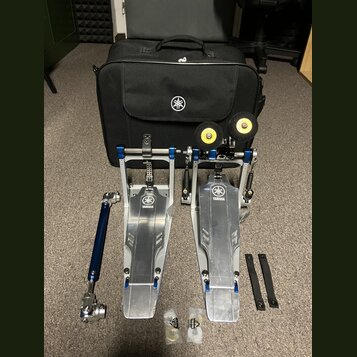 Sada kovových bubnových pedálů v modré a stříbrné barvě s logem YAMAHA. Součástí je černá přepravní taška s bílým logem Yamaha, dvě žluté gumové podl…