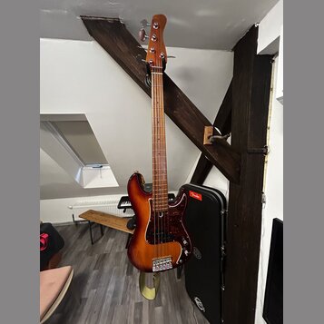 Elektrická basová kytara s trupem v červeně-žlutém sunburst provedení a tmavě červeným pickguardem. Na hlavě hmatníku není žádný viditelný nápis. V p…