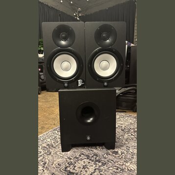 Sada tří černých studiových reproduktorů, dvě menší bedny umístěné nad jednou větší. Na spodní části každého reproduktoru je bílé logo Yamaha v kruhu…