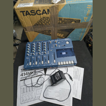 Modrý vícestopý magnetofon s nápisem TASCAM a označením PORTSTUDIO 414. Za přístrojem je umístěn originální karton s nápisy TASCAN, TASCAM a 4mkII. V…