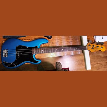 Modrá elektrická basová kytara s černým pickguardem a hnědým hmatníkem. Na hlavě nástroje je viditelné logo Fender a nápis PRECISION BASS. Nástroj vy…