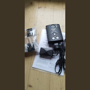 Na fotografii je černý ovládací modul značky Roland s tlačítky Tempo, Coach, Song, Drums a Select. Vedle něj leží černý pedál a uživatelská příručka…