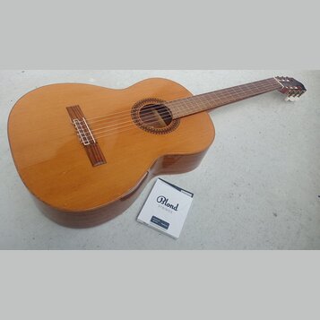 Kytara s přírodním dřevěným povrchem a hnědým hmatníkem. Vedle nástroje leží krabička s nápisy Blond STRINGS, classical guitar Medium a nečitelný tex…