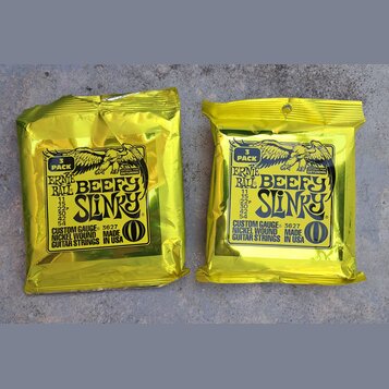 Dvě žluté fóliové balení kytarových strun. Na obou je nápis ERNIE BALL BEEFY SLINKY, CUSTOM GAUGE 3627, NICKEL WOUND MADE GUITAR STRINGS IN USA a ozn…