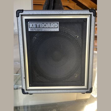 Kombinovaný zesilovač s šedým povrchem a černou síťovkou. V horní části je umístěn štítek s nápisy KEYBOARD a Roland SPECIALIZED MUSIC AMPLIFIER. Na…