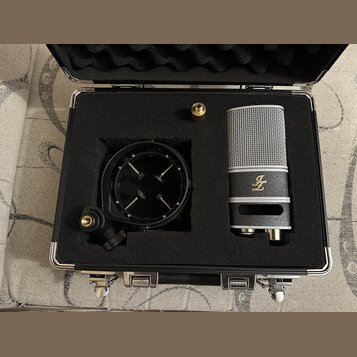 Kondenzátorový mikrofon stříbrné barvy s mřížkou a černý popfilter jsou umístěny v černé polstrované přepravní kufříku. Na těle mikrofonu je zlaté lo…