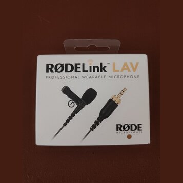 Fotografie znázorňuje bílé originální balení mikrofonu. Na krabici je uveden nápis RØDELink LAV a text PROFESSIONAL WEARABLE MICROPHONE, v pravém dol…