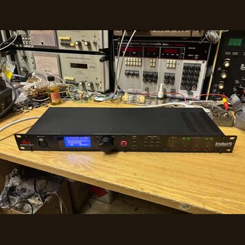 Černý rackový procesor s nápisem dbx PROFESSIONAL PRODUCTS a DriveRack PA2 SYSTEM MANAGEMENT SYSTEM. Zařízení má modrý podsvícený displej, ovládací k…