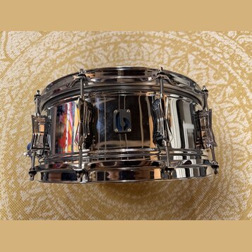Stříbrný chromovaný snare drum s kovovým korpusem a chromovaným kováním. Uprostřed korpusu je umístěn malý čtvercový štítek s modrým nápisem SONOR. P…