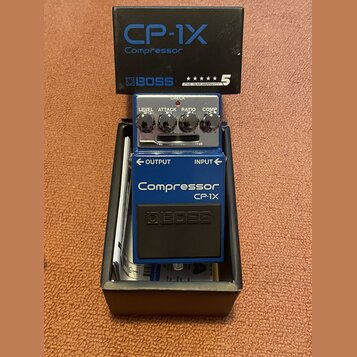 Modrý efekt pedál značky BOSS v originální krabici. Na pedálu jsou nápisy Compressor, CP-1X a čtyři ovládací prvky s popisky LEVEL, ATTACK, RATIO a C…