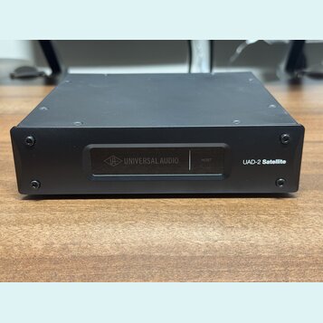 Černý kovový přístroj s hranatým tvarem. Na přední straně je nápis UAD-2 Satellite a displej s logem UA a textem UNIVERSAL AUDIO a HOST. Povrch je ma…