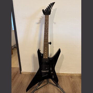 Černá elektrická kytara s ostře hranatým korpusem umístěná na stojanu. Na hlavě hmatníku je bílé logo Jackson. Nástroj má dva humbuckery, z nichž na…