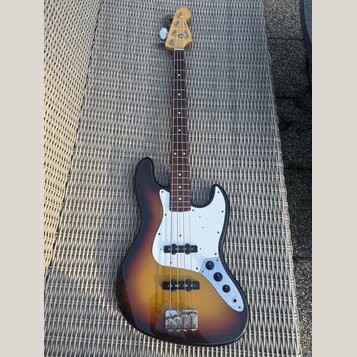 Čtyřstrunná elektrická basová kytara s tělem v provedení sunburst a bílým pickguardem. Na hlavě hředu je viditelné logo Fender. Nástroj vykazuje znám…