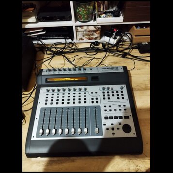 Černý kontrolér s kovovým horním panelem. V pravém horním rohu je nápis M-AUDIO PROJECTMIX I/O. Displej zobrazuje text M-Audio Control Surface is in…