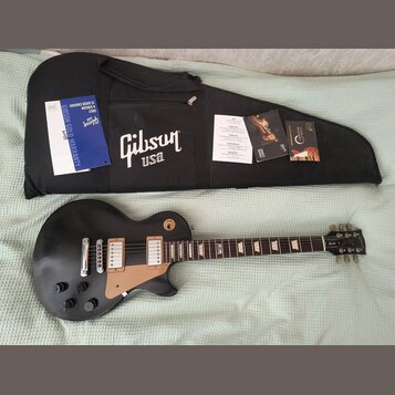 Černá elektrická kytara s hnědým pickguardem a dvěma humbuckery. Na hlavě nástroje je bílé logo Gibson. Vedle kytary je černý obal s bílým nápisem Gi…