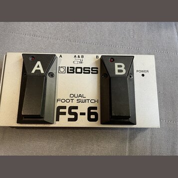 Stříbrný dvoupedálový spínací jednotka s černými plasty. Na přístroji jsou nápisy BOSS, DUAL FOOT SWITCH a FS-6 oraz označení pedálů A a B. V dolní č…