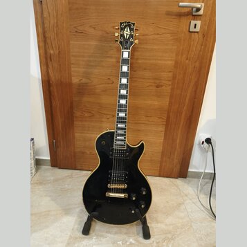 Černá elektrická kytara s gold hardwarem, umístěná na černém stojanu. Na hlavě je zlatý nápis Gibson. Nástroj má dva humbuckery, dva ovládací knofíky…