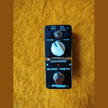 Černý kytarový efekt s nápisem BLACK TEETH a VINTAGE DISTORTION. Na horní straně jsou dva černé otočné knoflíky s popisky VOLUME a FILTER, jeden bílý…
