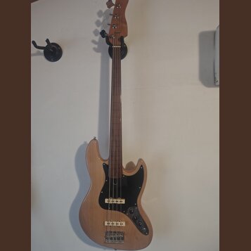 Elektrická basová kytara s přírodním dřevěným povrchem těla a hředu. Nástroj má černý pickguard, dva snímače a čtyři ladicí kolíky na hlavě. Na těle…