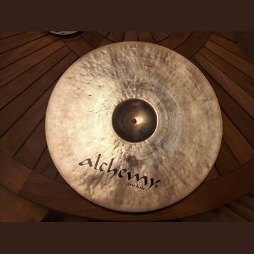 Kovový činel zlatavé barvy s viditelným rakováním povrchu. Na spodní části je černý nápis alchemy cymbals®. V horní části jsou drobné, lehce čitelné…