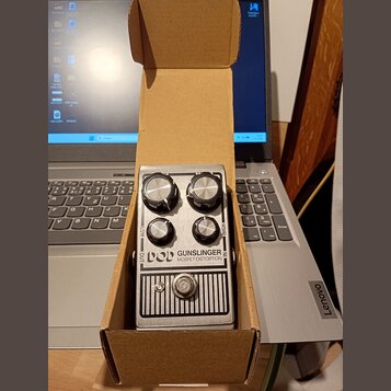 Stříbrný efekt pedál v kartonové krabici. Na přední straně je nápis POD GUNSLINGER MOSFET DISTORTION a čtyři ovládací knoflíky s popisky LOW, HIGH, O…