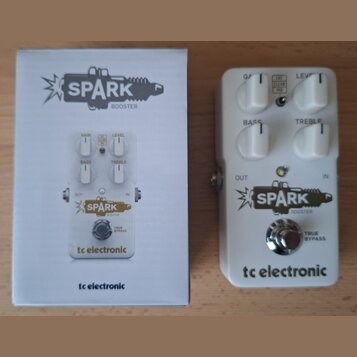 Bílá efekt pedál s nápisy SPARK BOOSTER a tc electronic. Pedál má čtyři bílé ovládací knoflíky s popisky GAIN, LEVEL, BASS a TREBLE, dále přepínač FA…