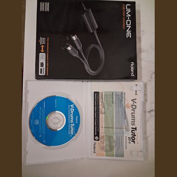Fotografie zobrazuje krabici s USB MIDI rozhraním Roland UM-ONE a otevřený plastový obal s CD diskem a příručkou. Na krabici je nápis UM-ONE USB MIDI…