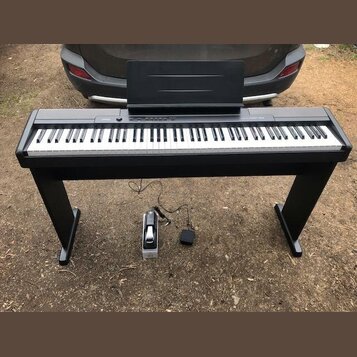 Černý digitální piano s klaviaturou a přiloženým notovým pultem na černém stojanu. Na pravé straně horního panelu je nápis CASIO PRIVIA. Pod nástroje…