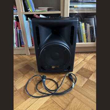 Černý kompaktní reproduktor s mřížkou a dvěma bočními rukojeťmi. Na spodní části mřížky je oranžové logo s nápisem soundnation. Před přístrojem leží…