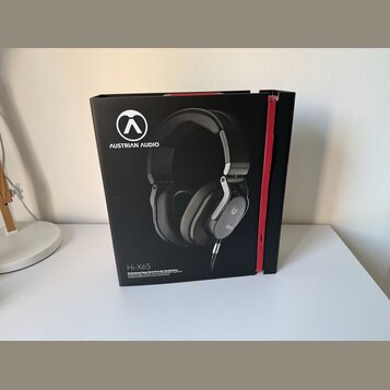 Černá kartonová krabice s červeným pásem na hraně. Na přední straně je fotografie černých sluchátek s logem a nápisem AUSTRIAN AUDIO. Dole je uveden…