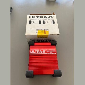 Červená DI box s hliníkovým žebrovaným povrchem a černými gumovými nohami. Na zařízení je bílý nápis ULTRA-G, logo BEHRINGER a text ACTIVE DI-BOX/VIR…