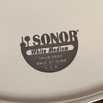Detail bílého bubnového koženě s černým potiskem. Je na něm vidět logo SONOR a nápisy White Medium, DRUM HEAD, MADE BY REMO a U. S. A. Povrch je hlad…