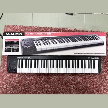 Fotografie zobrazuje černý klaviaturní kontrolér M-AUDIO KEYSTATION 61 MK3 a jeho původní kartonovou krabici. Na přístroji i na obalu je vidět značka…